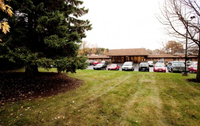 A small parking lot with several cars in front of a building surrounded by grass and trees.