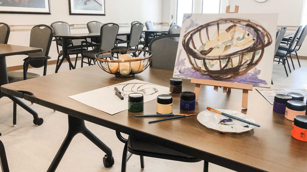 A painting setup featuring a canvas on an easel and various art supplies, with a still life composition of fruit in a basket.