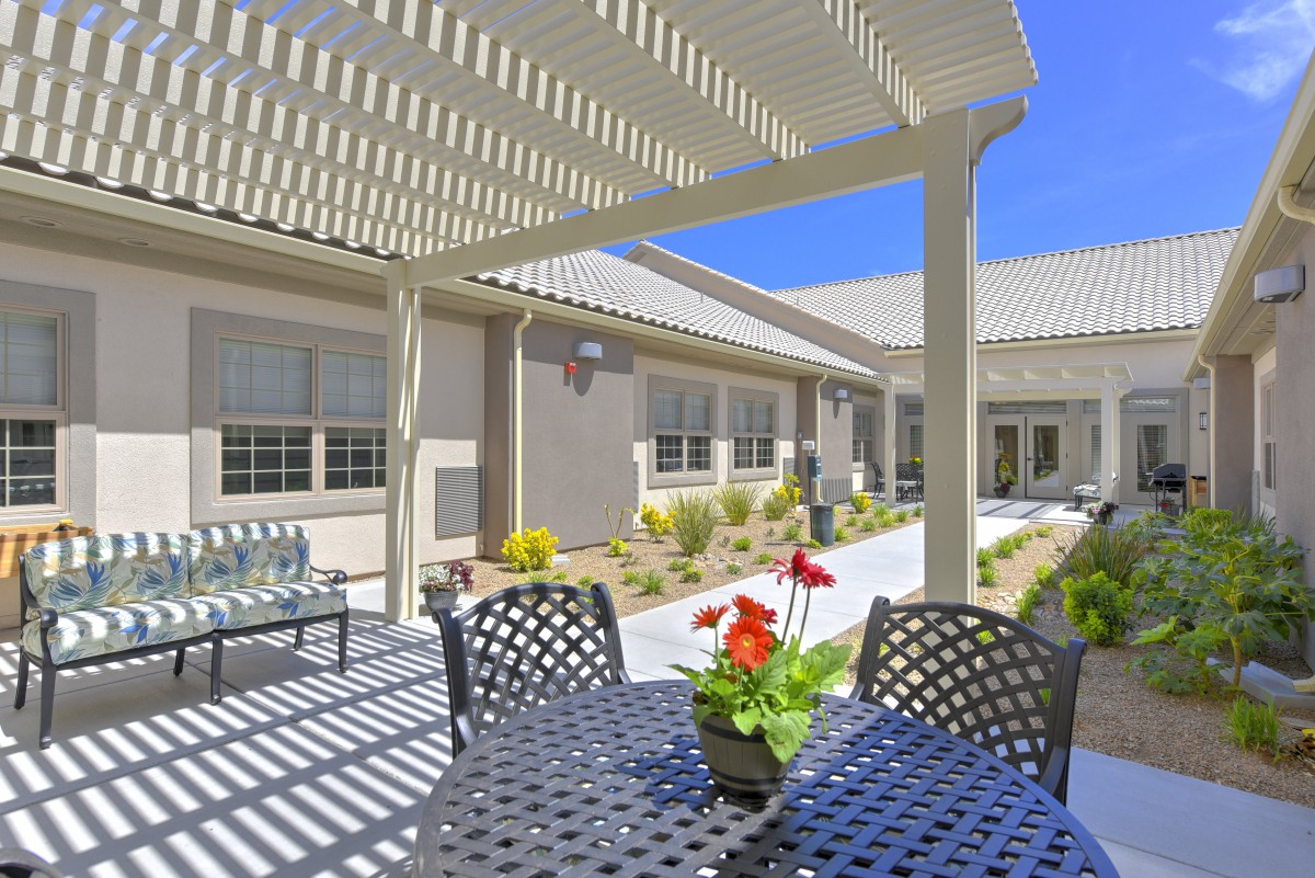 A sunny outdoor patio with a pergola providing shade over a table and chairs, adorned with red flowers.