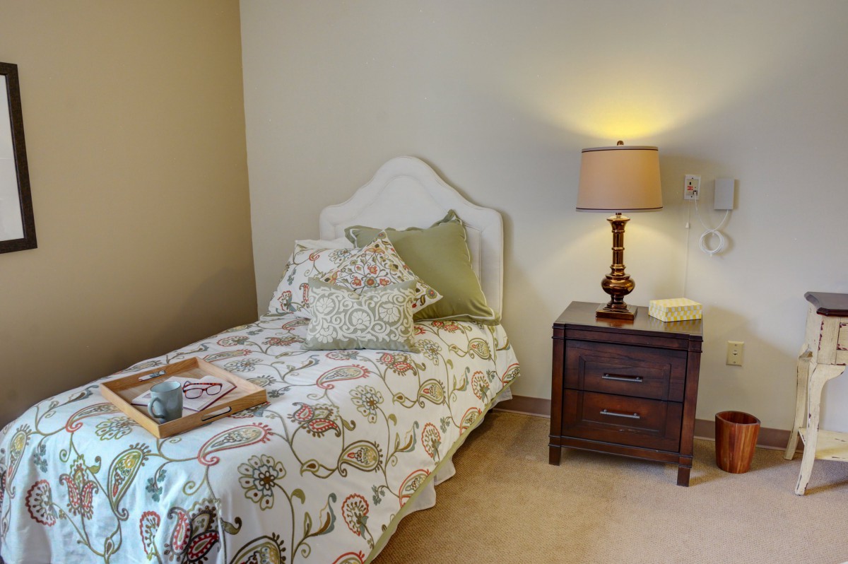 A cozy bedroom featuring a single bed with decorative bedding, a nightstand, and a lamp.
