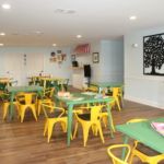 A bright and colorful children's classroom with green tables and yellow chairs.