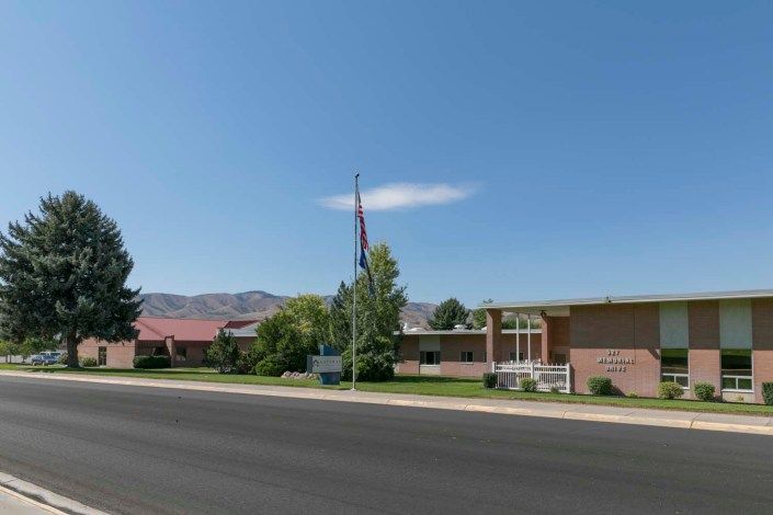 A building with a flagpole in front, set against a backdrop of mountains and trees under a clear sky.