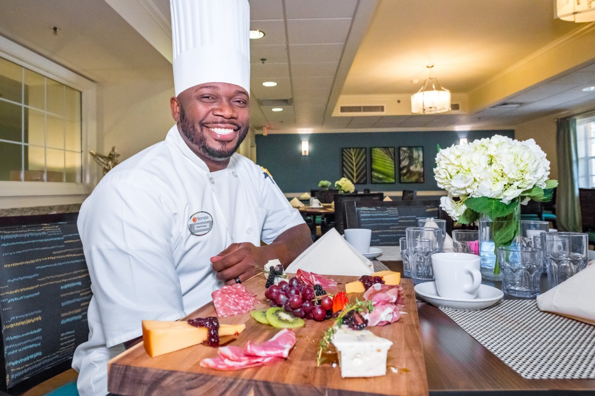 A smiling chef presents a beautifully arranged charcuterie board with various cheeses and fruits in a restaurant setting.