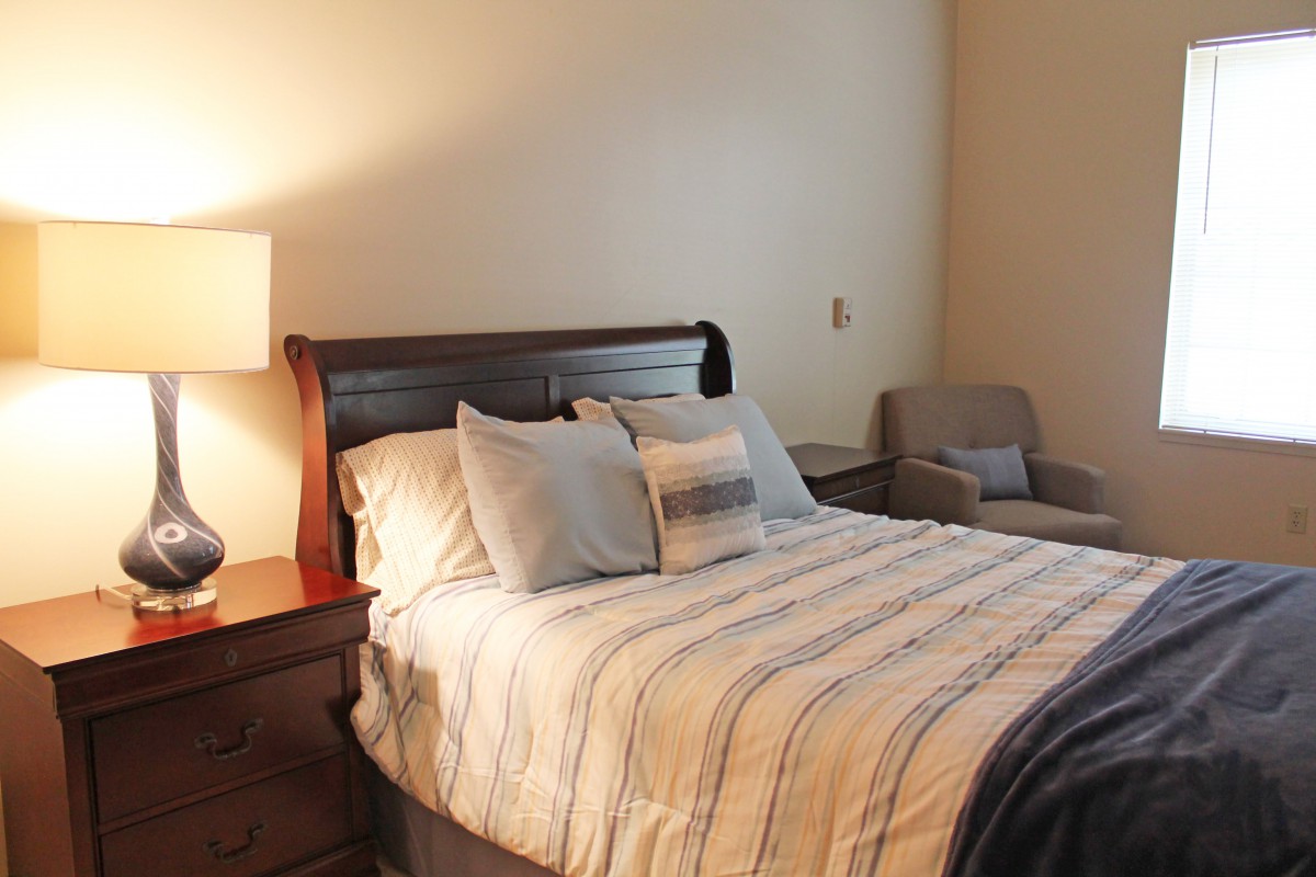 A cozy bedroom with a neatly made bed, an armchair, and a bedside lamp.