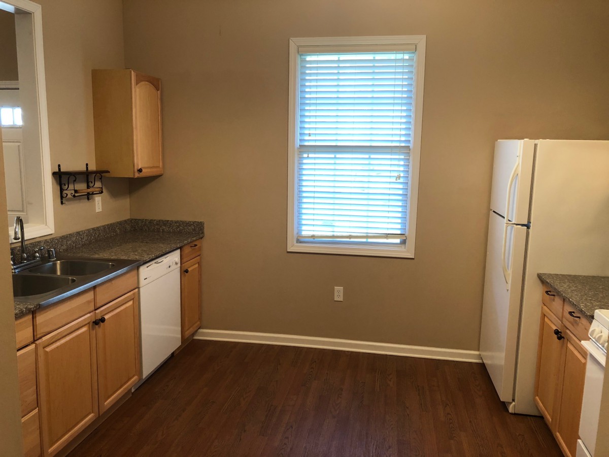 An empty kitchen with wooden cabinets, a refrigerator, dishwasher, and a window with blinds.