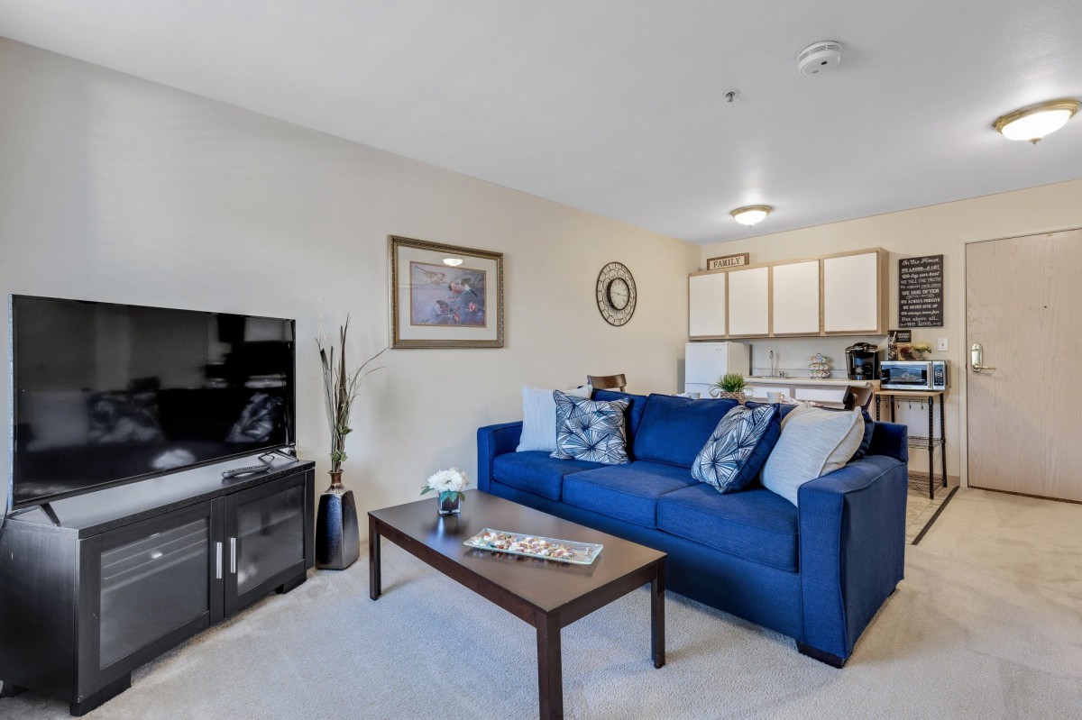 A cozy living room with a blue sofa, coffee table, and TV, connected to a kitchen area.