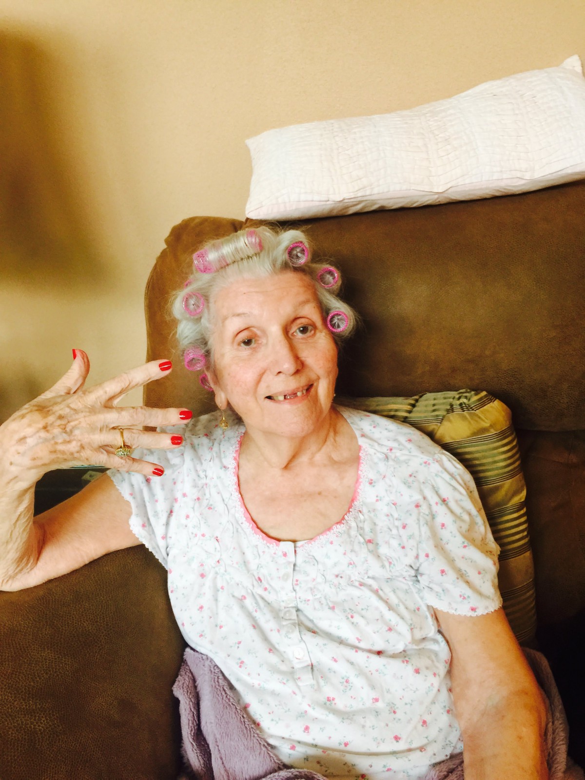 An elderly woman sits comfortably with hair in curlers and freshly painted nails, smiling warmly at the camera.