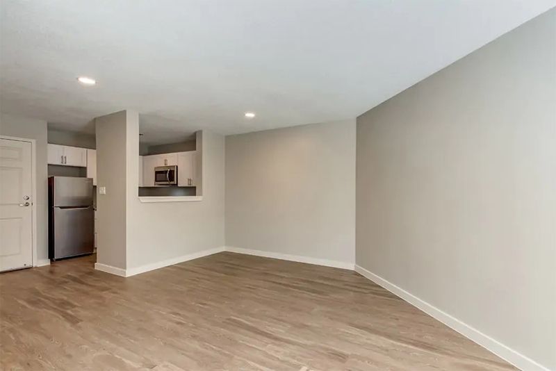 An empty room with wood flooring and a view into the kitchen, featuring an open floor plan and recessed lighting.