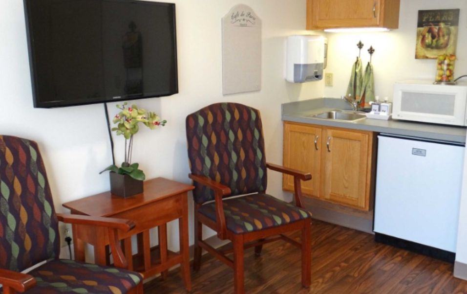 A cozy living space with a patterned armchair, wooden table, wall-mounted television, and a small kitchenette.