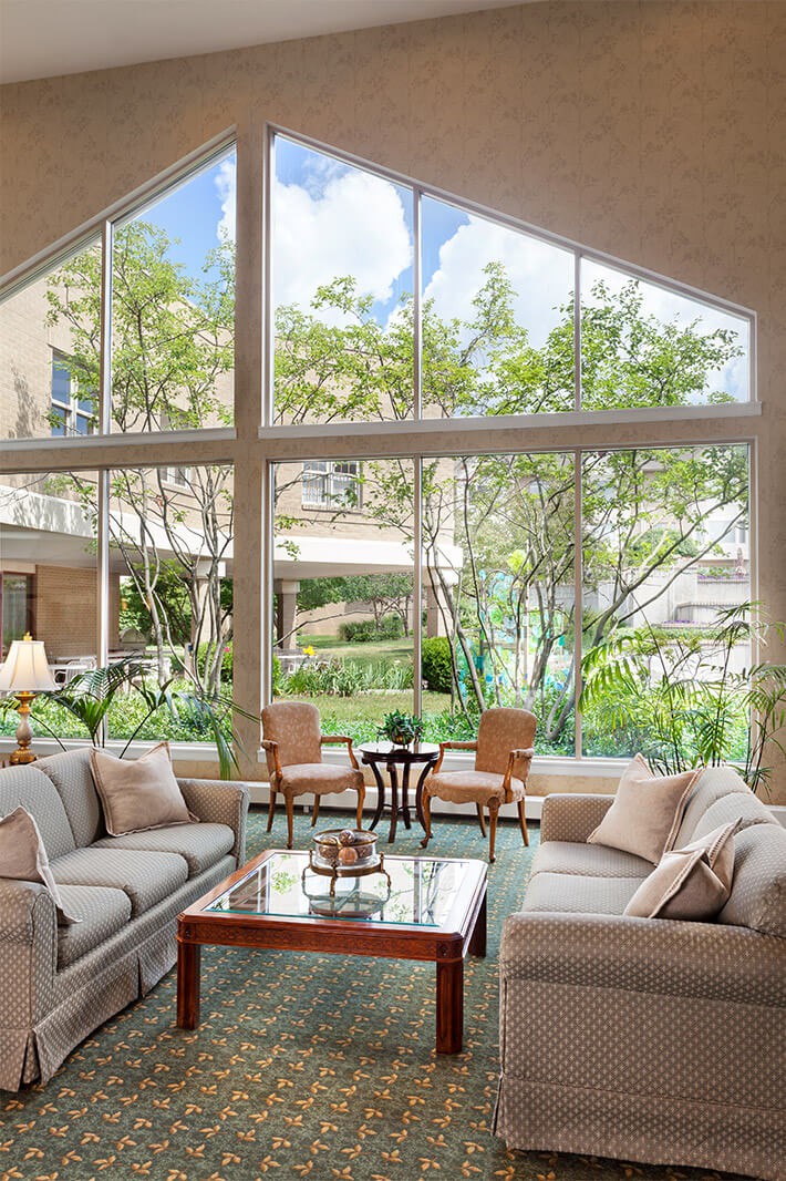 A spacious living room with large windows allowing ample natural light, featuring comfortable sofas and a glass coffee table surrounded by greenery.