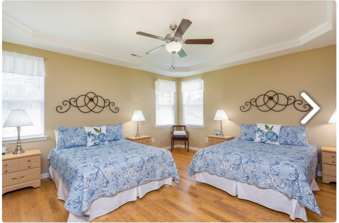 A bright bedroom featuring two twin beds with blue bedding, a ceiling fan, and elegant decor.
