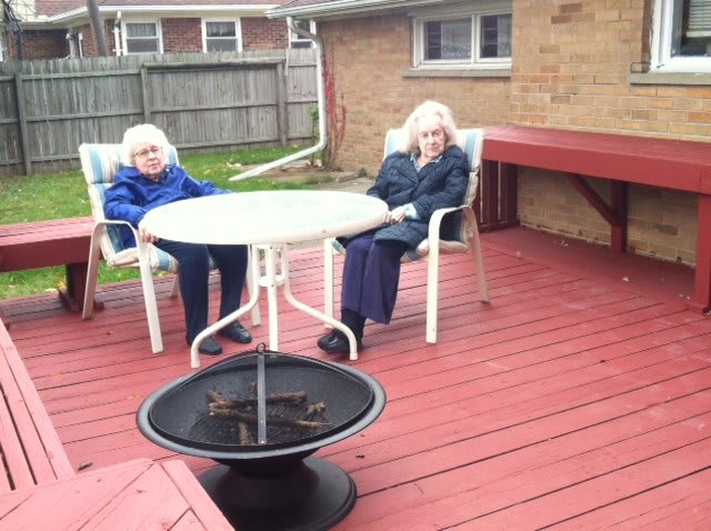 Two elderly individuals sitting on a wooden deck with patio furniture and a fire pit.