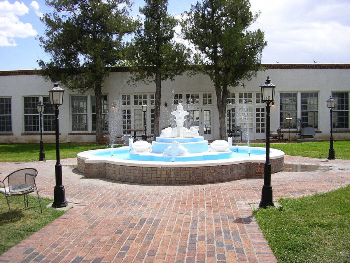 A tranquil courtyard with a central fountain, surrounded by lampposts and trees, adjacent to a building.
