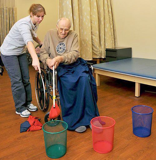 An elderly person in a wheelchair is assisted by a caregiver during a physical therapy session involving colorful baskets and bean bags.