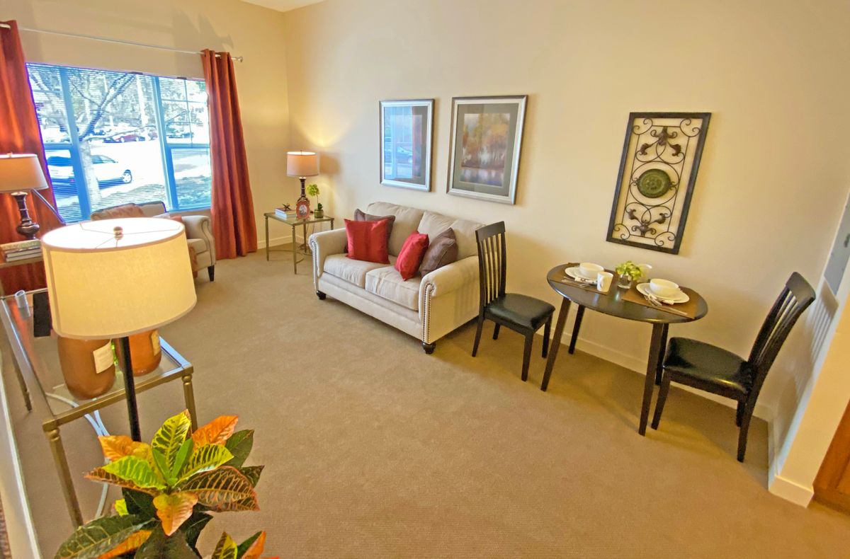 A cozy living room with a sofa, decorative pillows, and artworks on the wall. There's a small dining table with two chairs and a window with red curtains providing natural light.