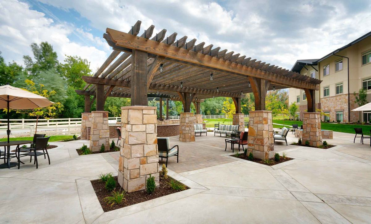 A well-designed outdoor pergola with stone pillars, featuring tables and chairs in a landscaped park setting.