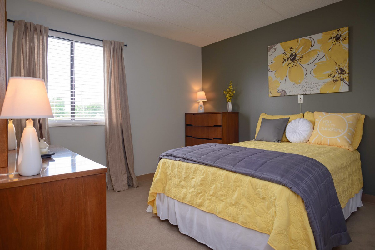 A modern bedroom featuring yellow bedding and a mix of neutral tones, with a dresser, lamp, and art on the walls.