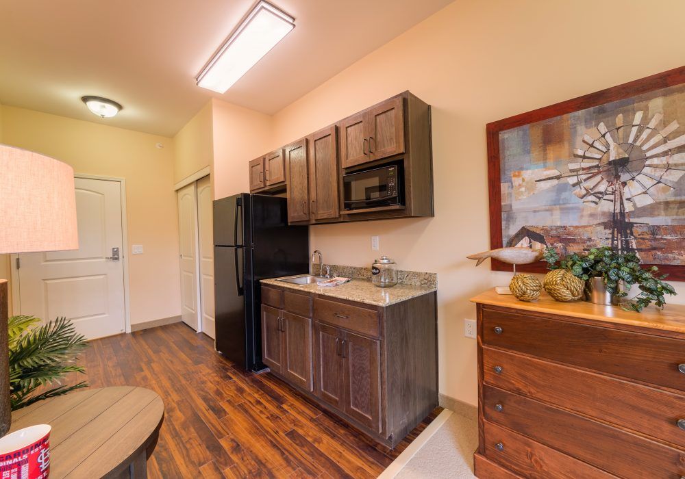 A compact kitchen with dark wood cabinets, a black refrigerator and microwave, and wooden flooring.