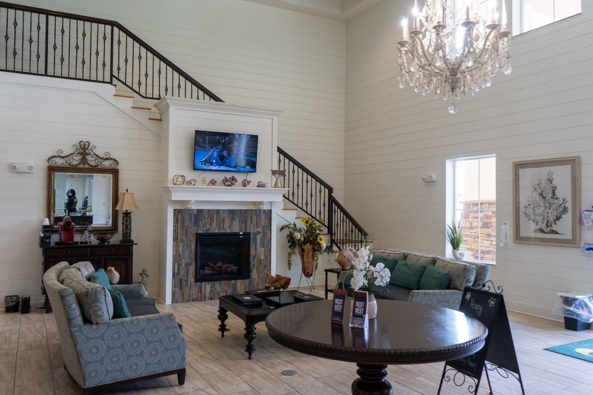 A spacious living room with high ceilings, featuring a grand chandelier, a television mounted above the fireplace, and comfortable seating around a coffee table.