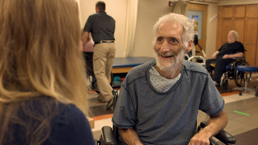 An elderly man smiling during a conversation with a caregiver in a rehabilitation setting.