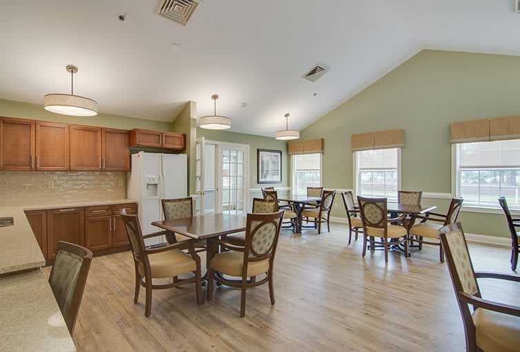 A spacious dining area with multiple tables and chairs, adjacent to a kitchen, featuring wooden floors and bright lighting.