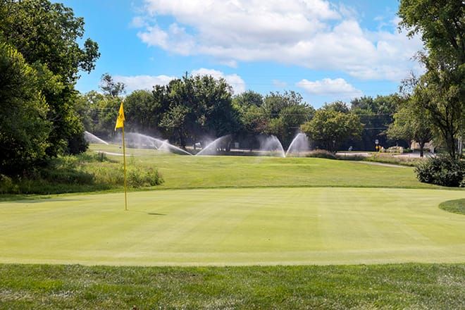 A well-maintained golf course featuring a green with sprinklers in operation under a clear blue sky.