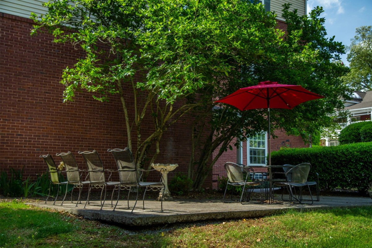 A cozy outdoor patio with chairs and a table under a red umbrella, set against a brick wall and shaded by a lush tree.
