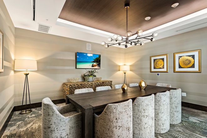 A modern dining room featuring a large table with upholstered chairs, a stylish chandelier, and decorative elements including wall art and a television.