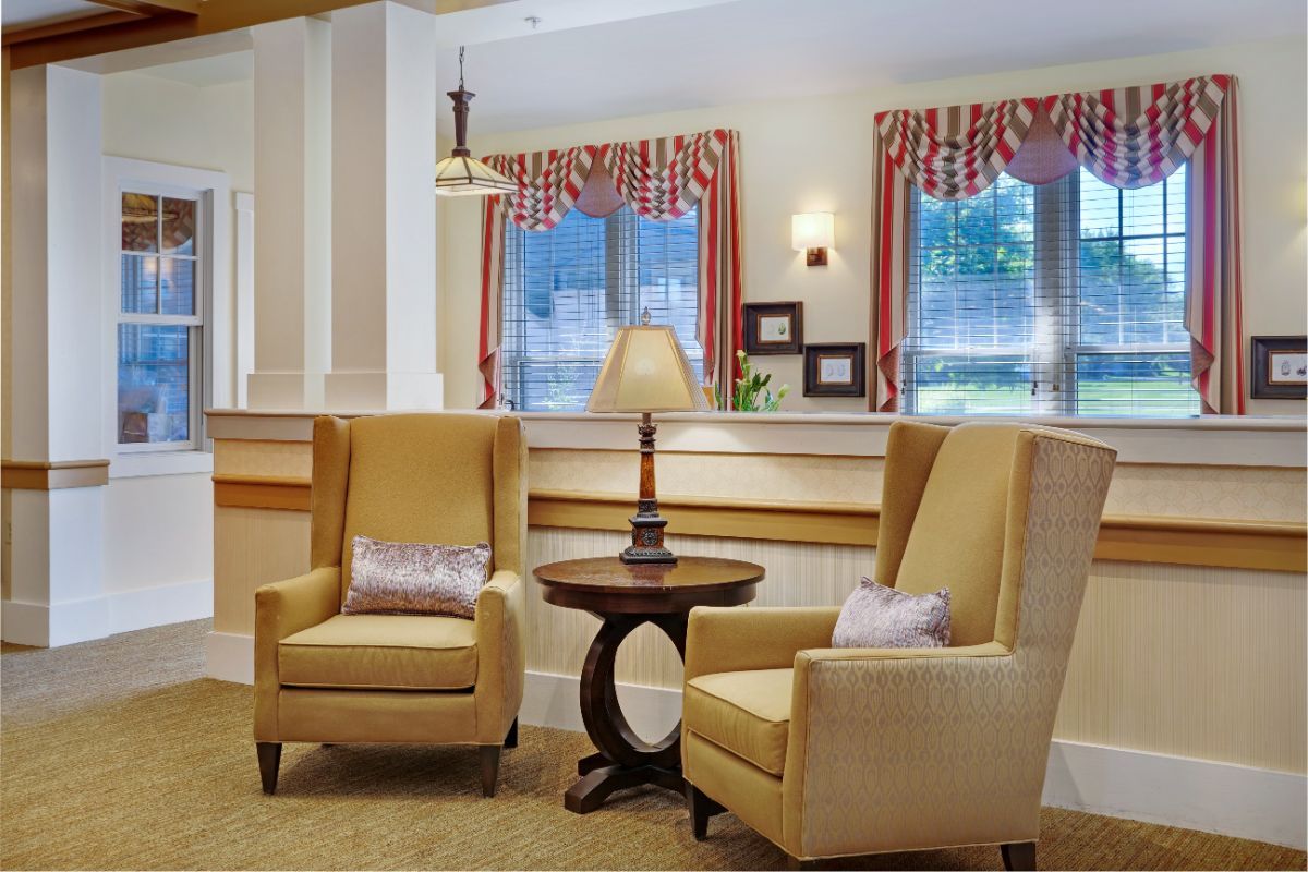 A cozy lounge area with two armchairs and a table topped with a lamp, framed by windows with red and white curtains.