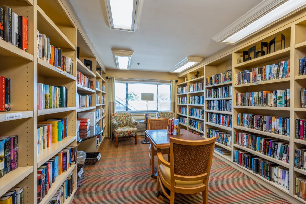 A cozy library room with bookshelves lining the walls, a table and chairs in the center, and a window providing natural light.