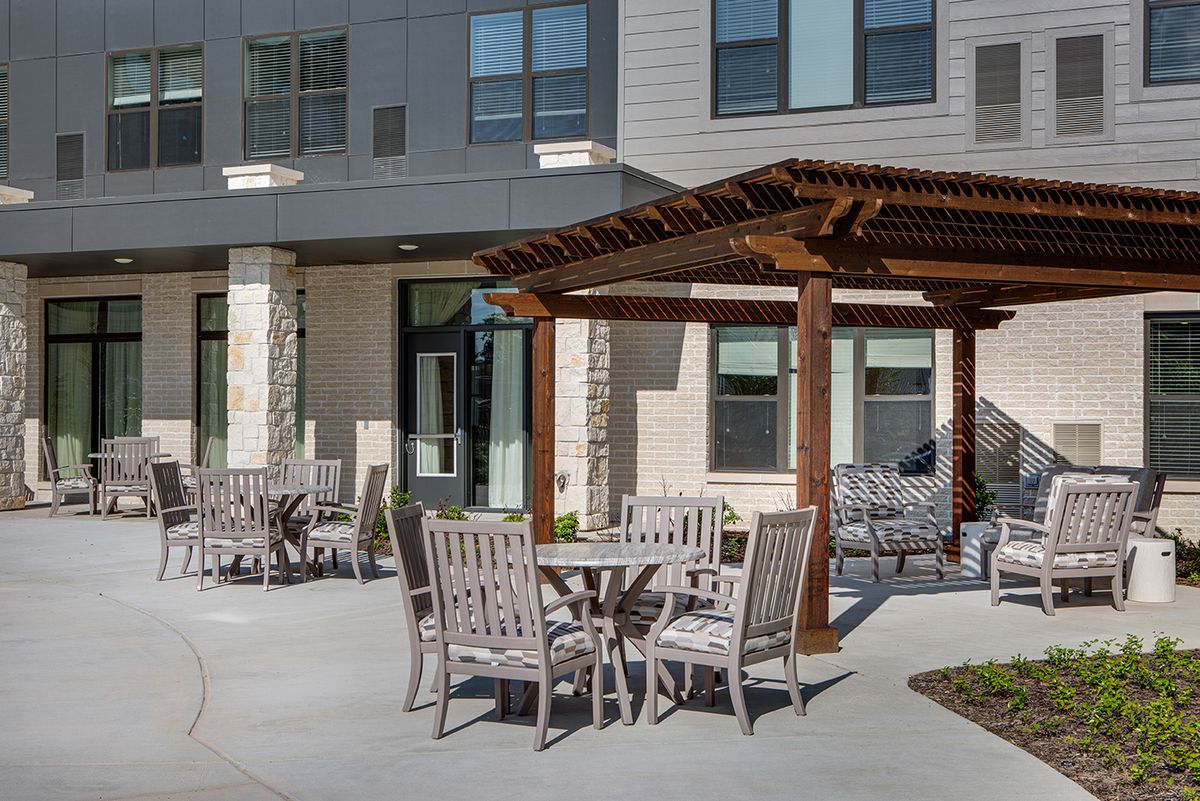 A well-furnished patio area with a pergola, featuring wooden tables and chairs outside a modern building.