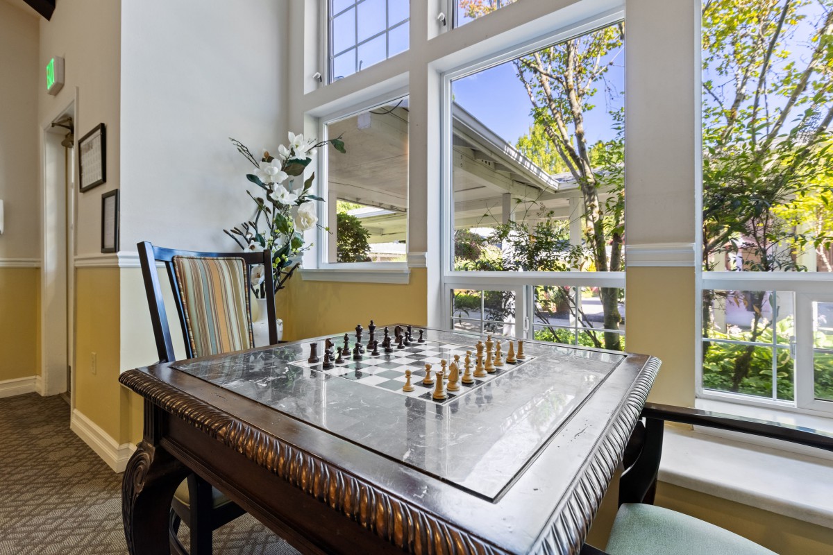 A chessboard set up on a game table near a large window with an outside view and natural lighting.