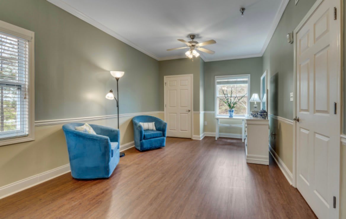 A cozy living room with blue chairs, a standing lamp, and a ceiling fan, featuring hardwood floors and natural light from a window.