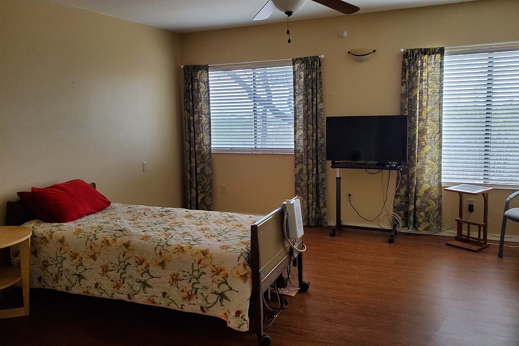 A cozy bedroom with a floral-patterned bed, window draped with curtains, and a TV on a stand.