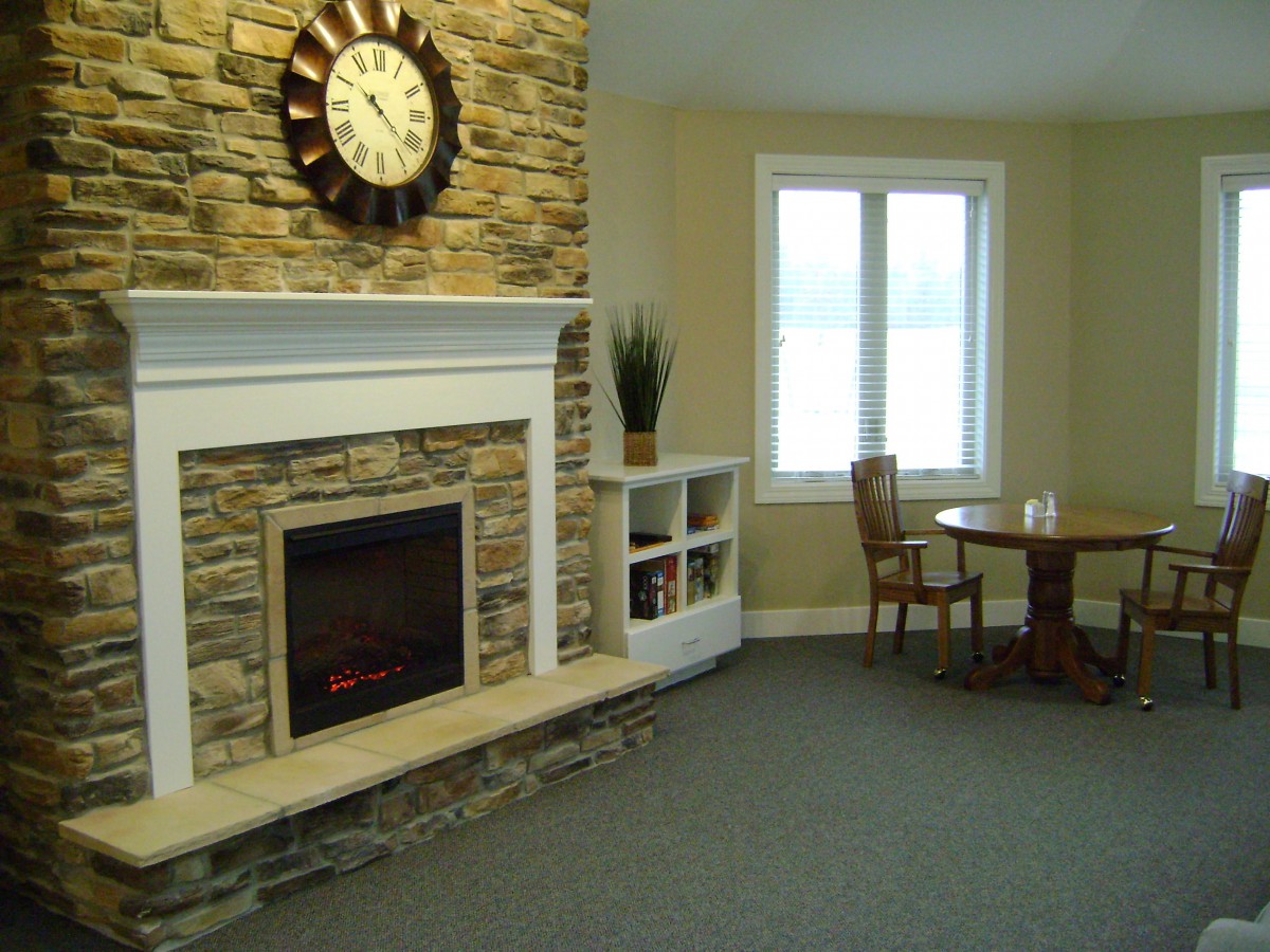 A cozy living room featuring a stone fireplace with a clock above it, alongside a small table and chairs near a window.