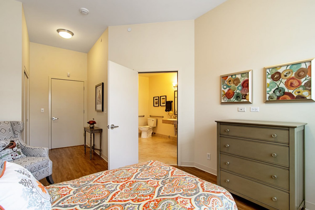 A cozy bedroom with a patterned bedspread, a dresser, and an open doorway leading to a bathroom.