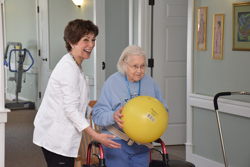 A caregiver joyfully assists an elderly woman as she engages in a fitness activity using a yellow ball.