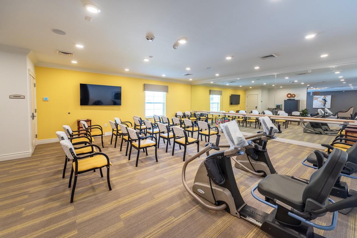 A modern exercise room features rows of chairs, fitness equipment, and a television mounted on a yellow wall.