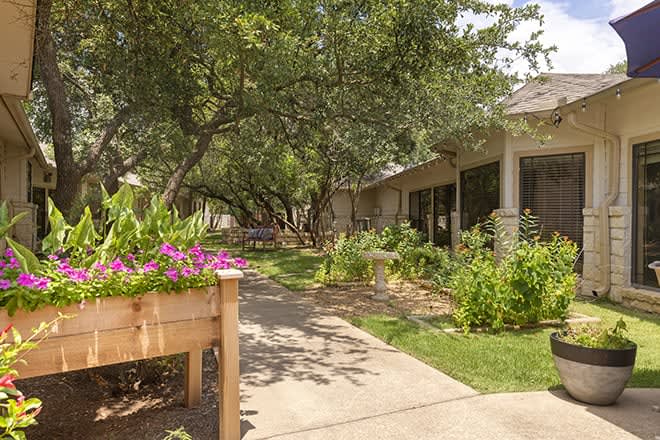 A peaceful garden with a pathway leading to a house, surrounded by trees and vibrant flowers.