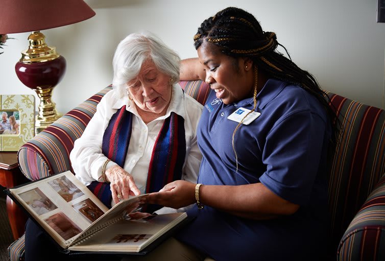 A caregiver and an elderly woman look through a photo album together in a cozy home setting.
