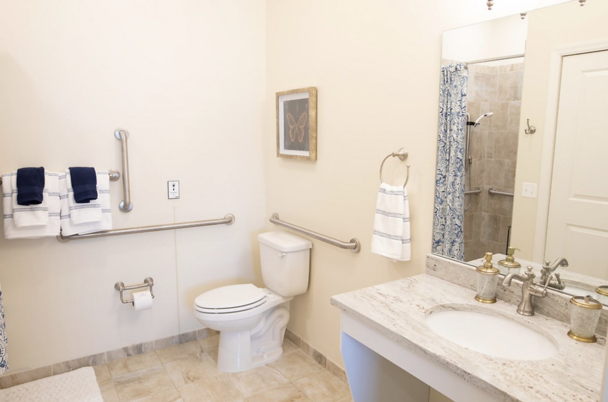 An accessible bathroom featuring a toilet with handrails, a sink with a mirror above, and a towel rack.