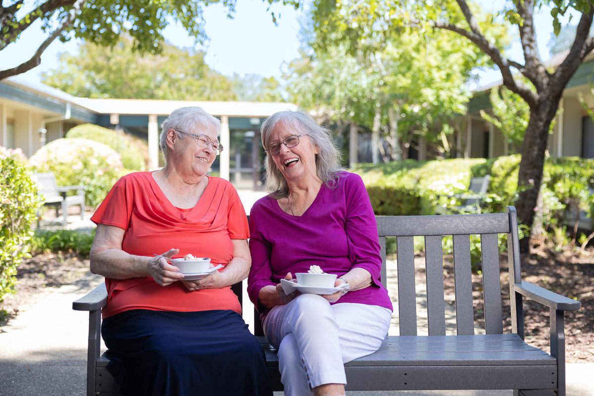 Two elderly women enjoying a conversation and tea together on a bench outdoors.