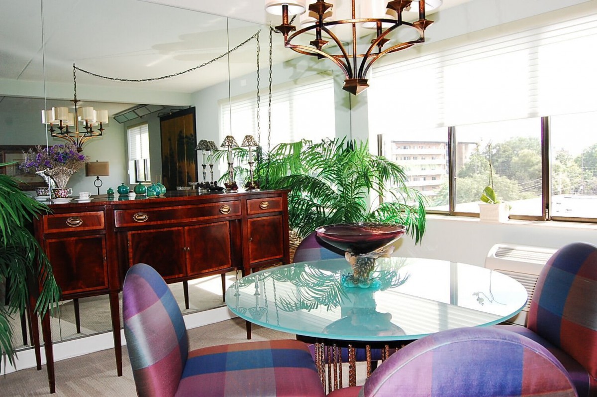 A well-lit dining room with a glass table and elegant chairs, featuring a large mirror and decorative plants.
