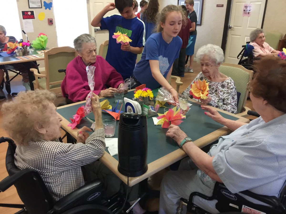 A group of elderly women engage in a crafts session with younger volunteers at a nursing home.