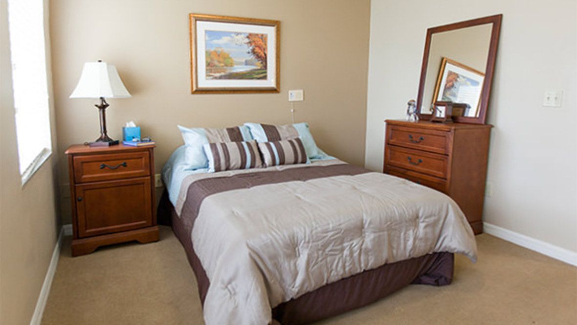 A cozy bedroom featuring a neatly made bed with pillows and a comforter, accompanied by a nightstand with a lamp, a dresser with a mirror, and a landscape painting on the wall.