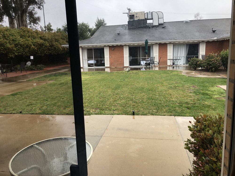 A rainy day view of a backyard with a lawn and patio area facing a house.