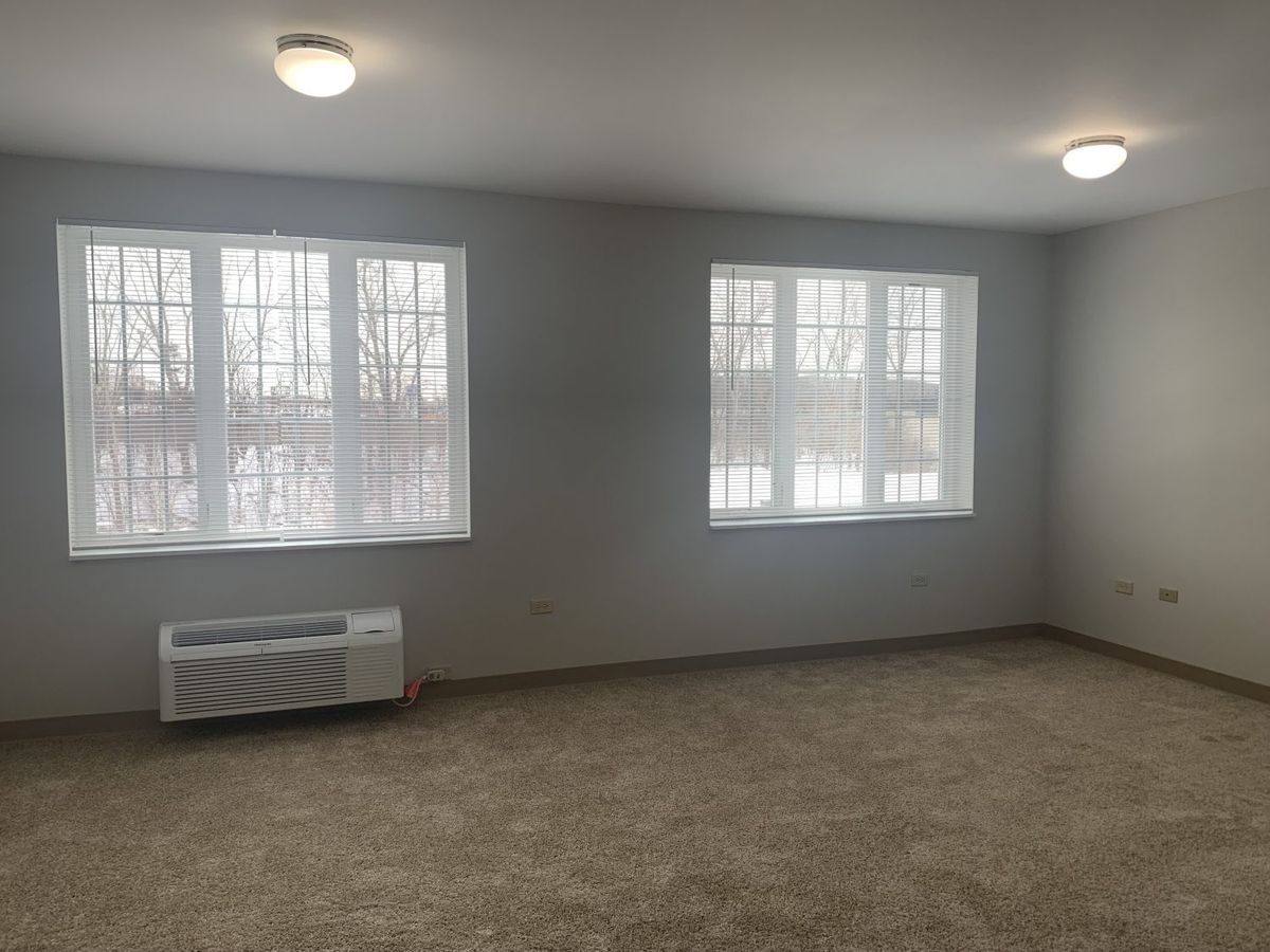 An empty room with carpet flooring, two windows, and ceiling lights.