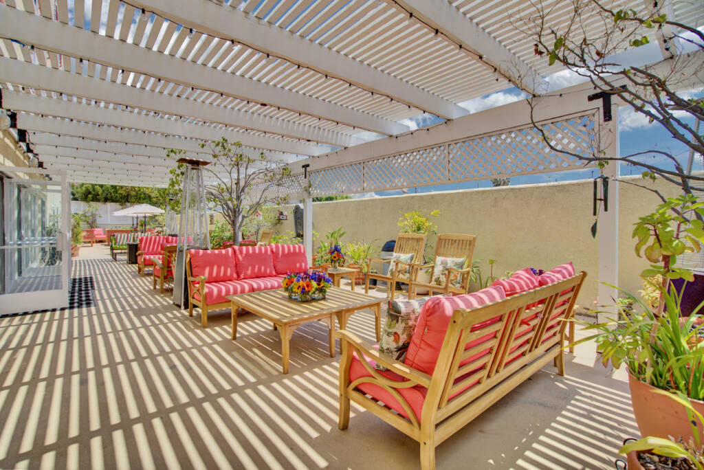 A sunlit patio with wooden furniture and red cushions under a pergola.