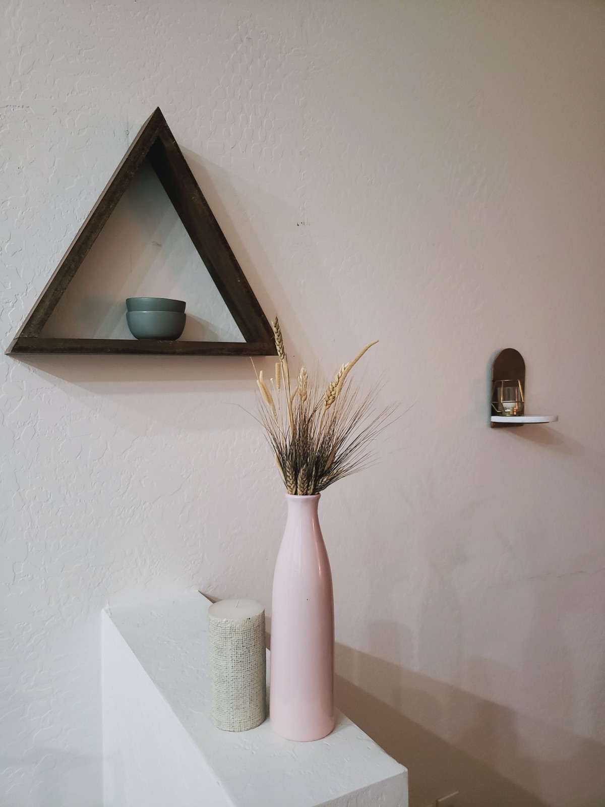 A minimalist interior featuring a wooden triangle shelf with a ceramic bowl, alongside a pink vase filled with dry grass.