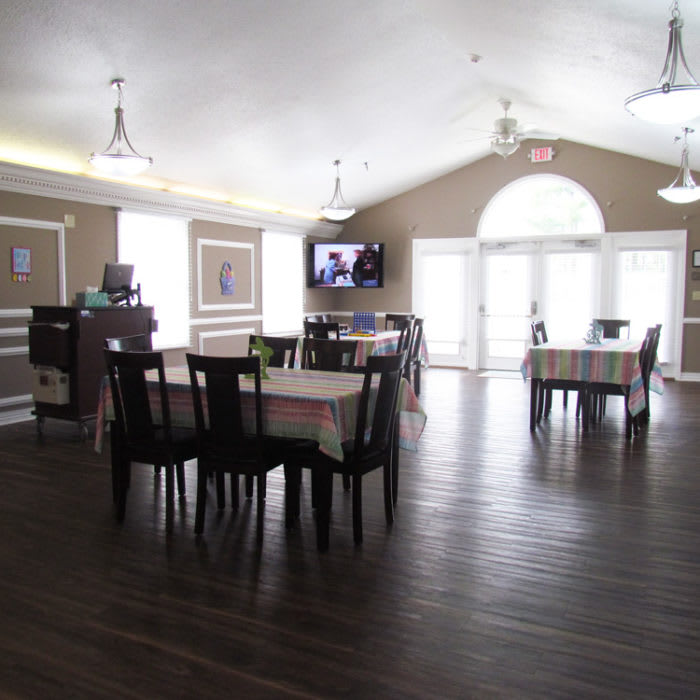 A spacious dining area with tables, chairs, and a television, featuring large windows and a wooden floor.